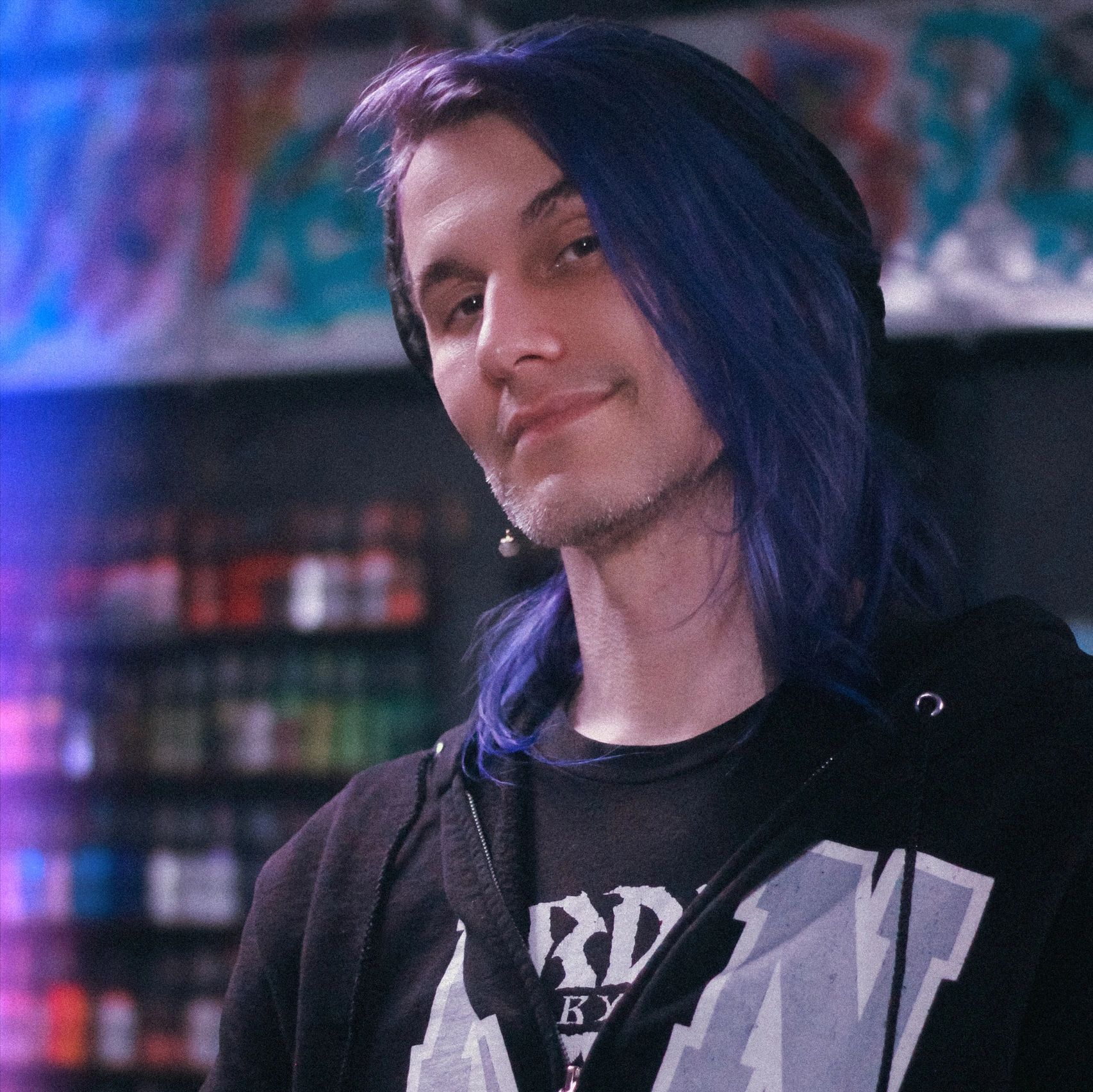 Person with long purple hair smiling in a dimly lit room with colorful background.