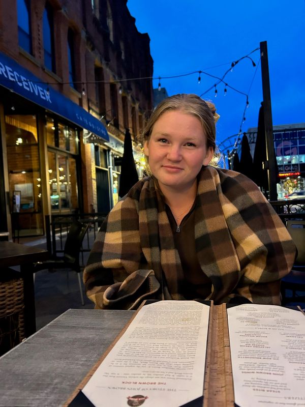 Woman wrapped in a checkered blanket at an outdoor restaurant at dusk.