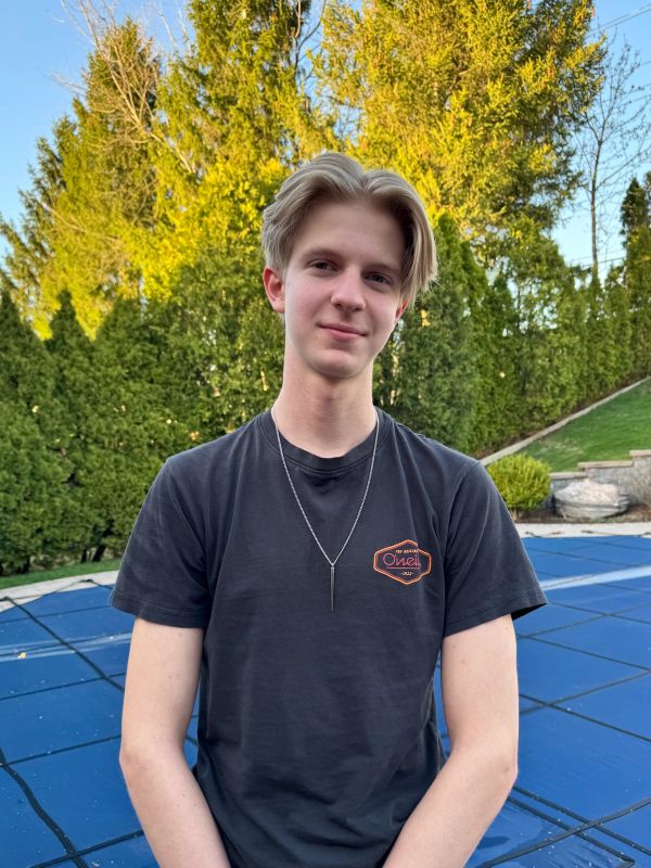 A young man in a black t-shirt standing outdoors with greenery in the background.