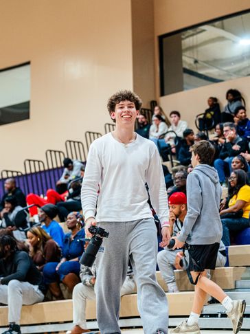 Smiling young man holding a camera at a crowded indoor event.