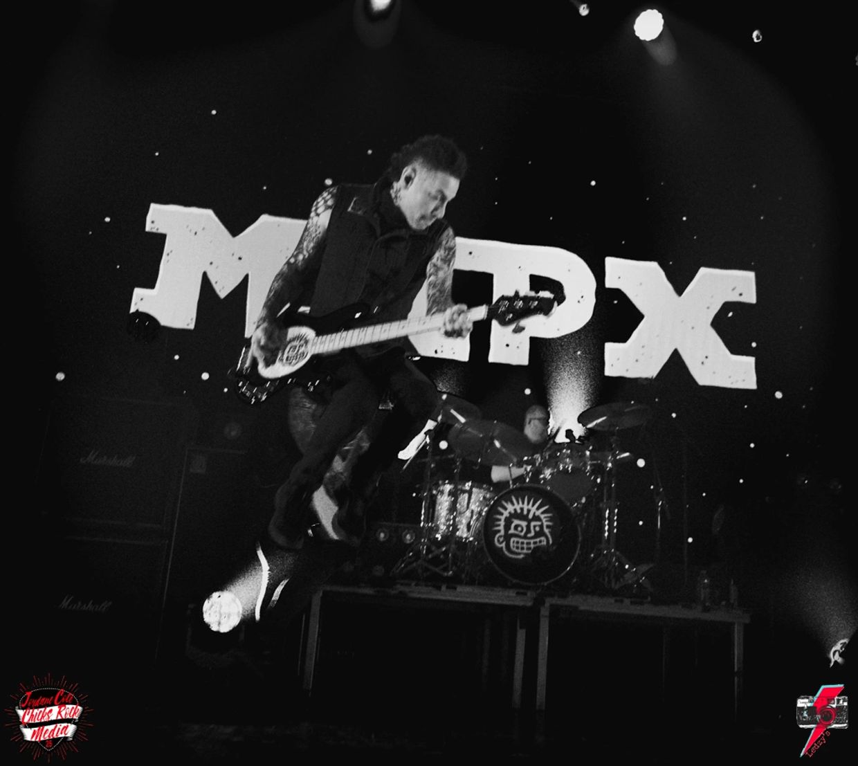 Bassist jumps energetically on stage during a live concert with drummer in the background.