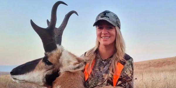 A woman in camouflage gear posing with a pronghorn antelope in a field.