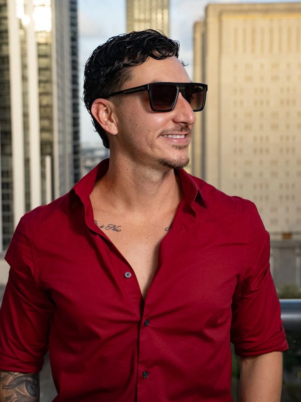 Man in a red shirt and sunglasses smiling outdoors in an urban setting.