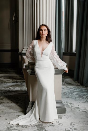 Woman in elegant white gown with sheer floral sleeves poses confidently indoors.