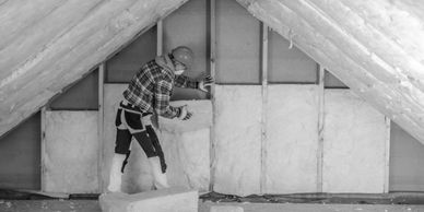 Worker installing fiberglass insulation in attic space with safety gear.