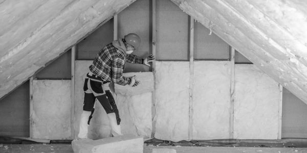 Worker installing fiberglass insulation in attic space with safety gear.