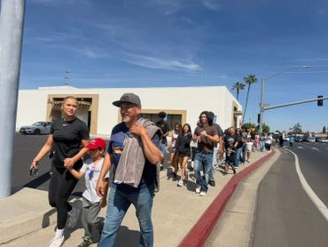A diverse group walking along a sunny sidewalk near an intersection.