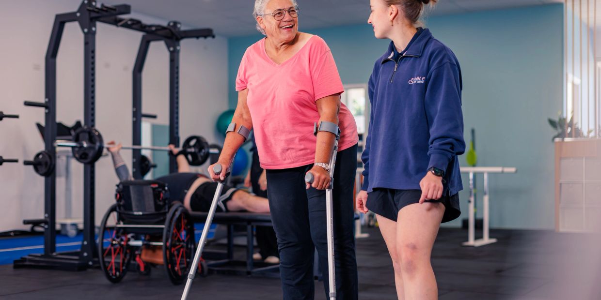 Clinician walks with smiling older woman using crutches through accessible gym