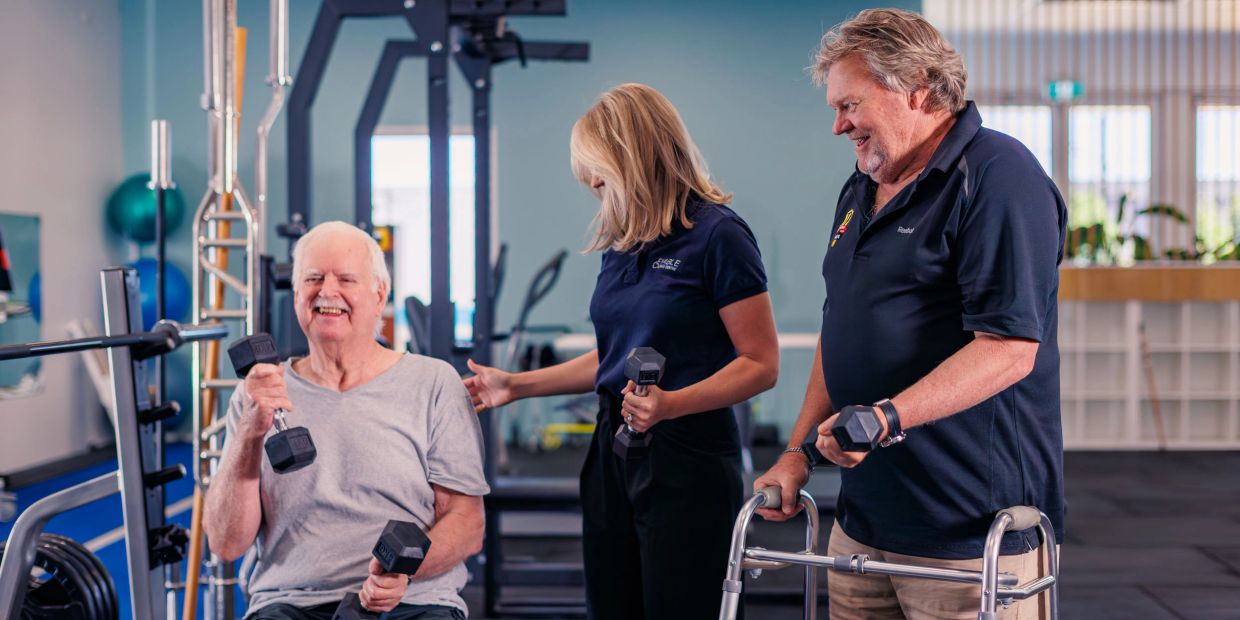 A female clinician supporting two older, smiling men to lift weights in a gym.