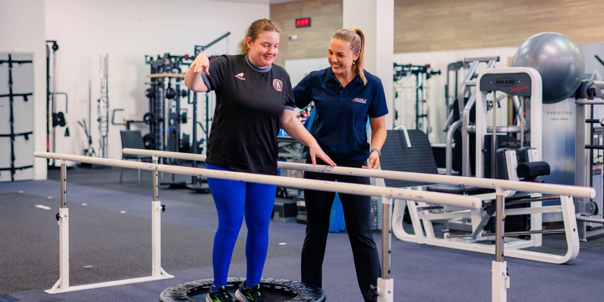 Clinician assisting a woman with a disability with balance exercises on a trampoline at a gym.