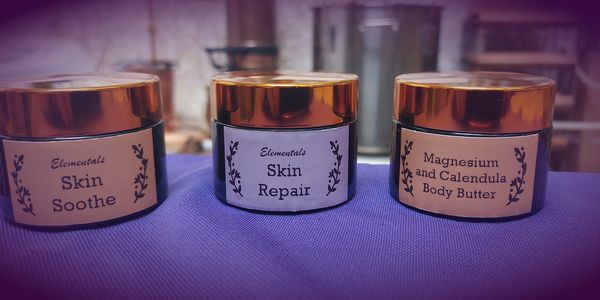 Three jars of Elementals skin care products with gold lids on a purple cloth.