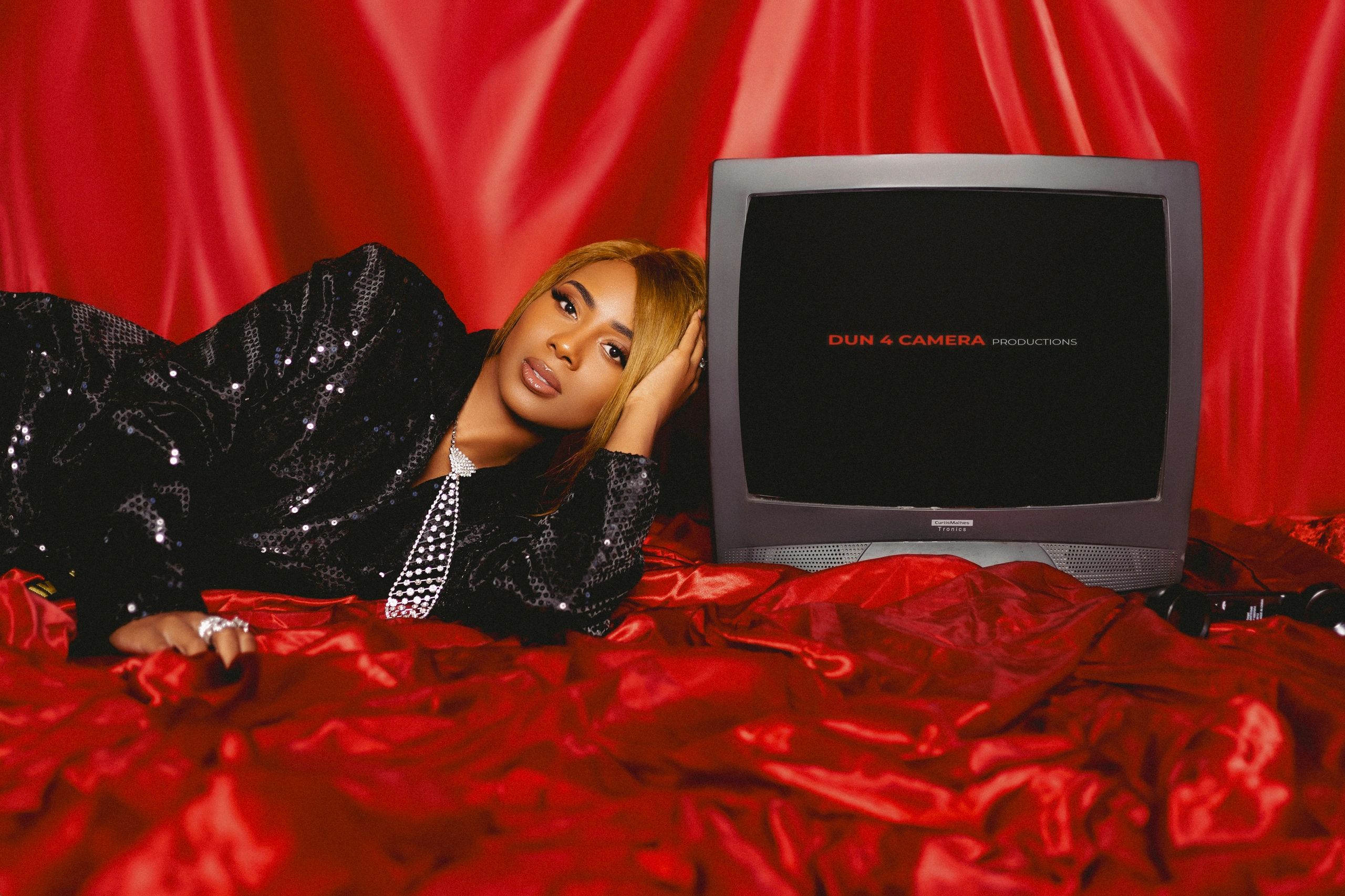 Woman in black sequin outfit lying on red satin beside an old TV.