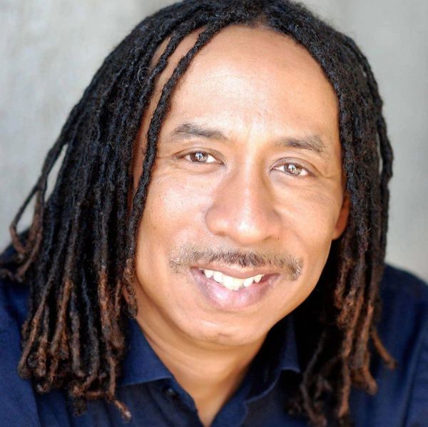 Smiling man with dreadlocks wearing a dark shirt.