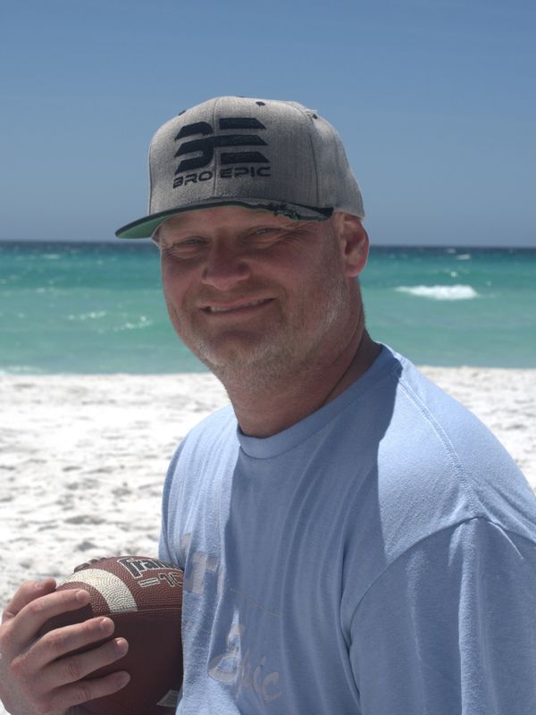 Man in a gray cap holding a football at the beach on a sunny day.