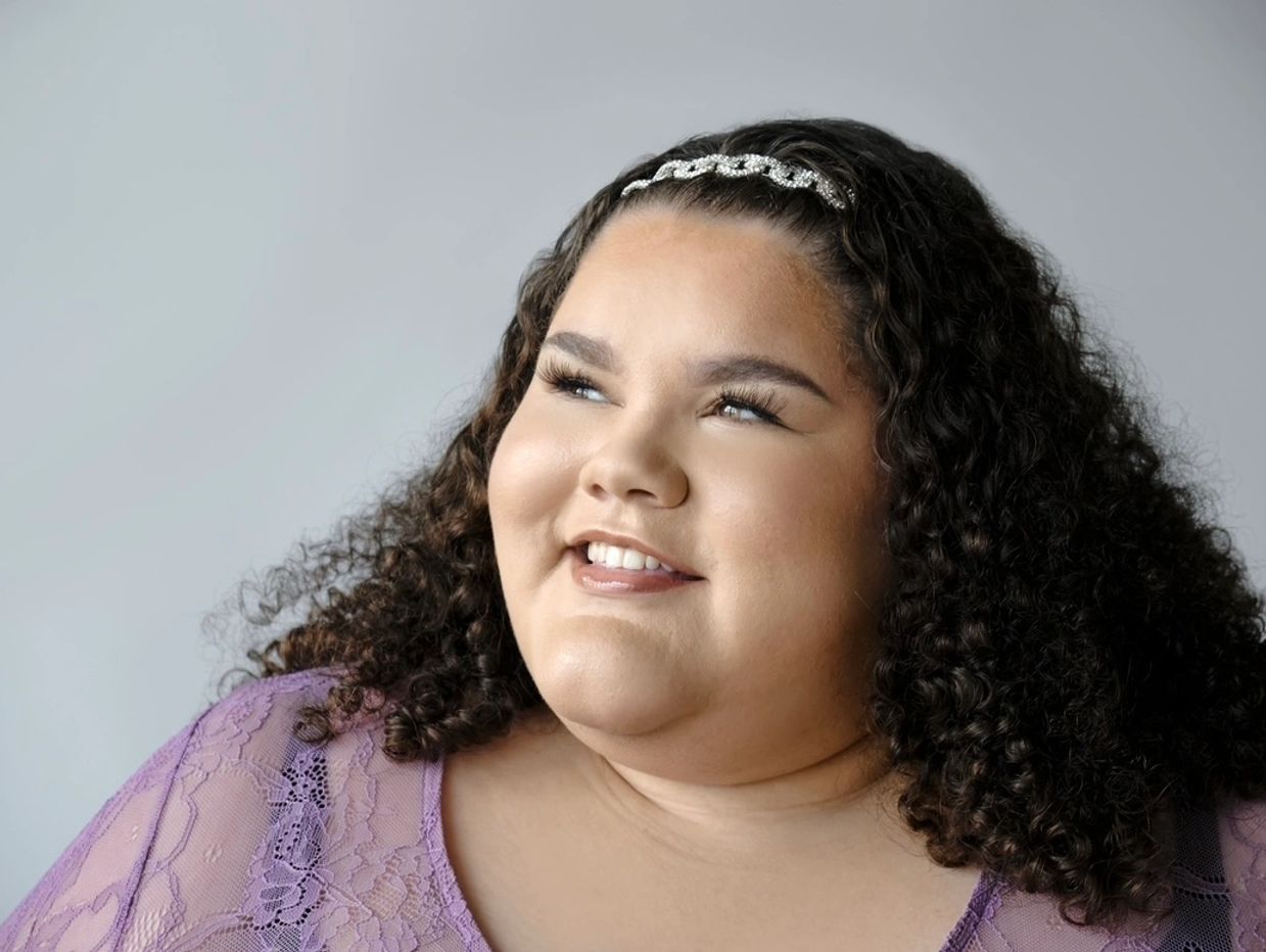 Plus Size Travel Expert Jaelynn Chaney is sitting while smiling off to the side in a purple lace top