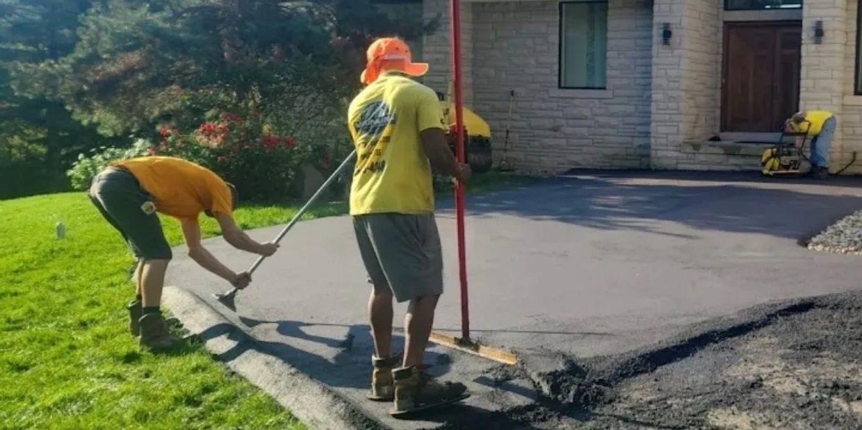 Two workers smoothing freshly laid asphalt on a driveway in front of a house.