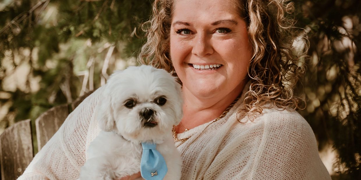 Smiling woman holding a small white dog with a blue tie outdoors.