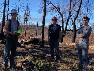 Three smiling young men in the forest, wearing hard hats and holding tree-falling wedges. 