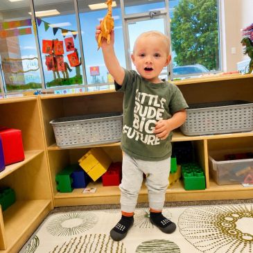 Toddler holding up a toy dinosaur in a colorful playroom.