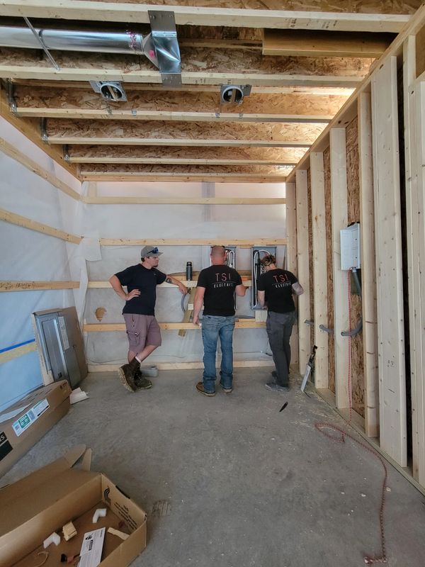 Three electricians working on wiring inside a wooden-framed construction site.