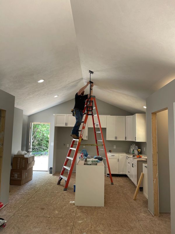 A worker installs a ceiling fixture on a ladder in a kitchen under renovation.
