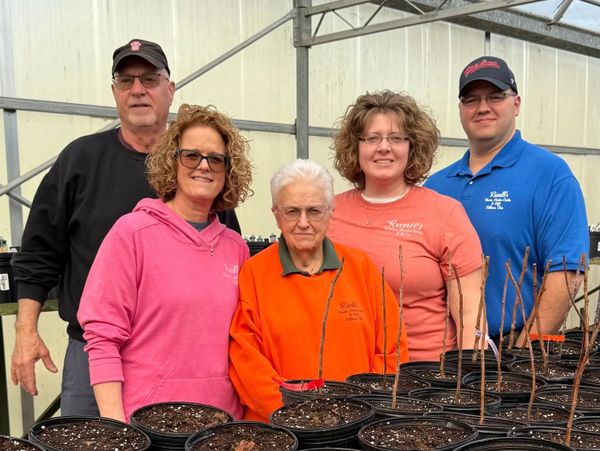 Five people standing behind rows of black plant pots in a greenhouse.