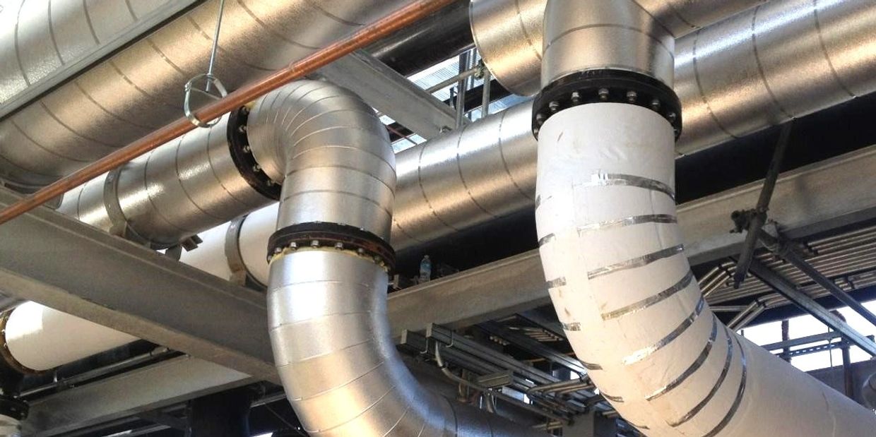 Large metallic and insulated pipes in an industrial facility ceiling.