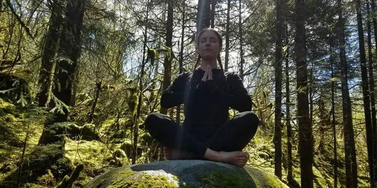 Person meditating on a mossy rock in a sunlit forest.