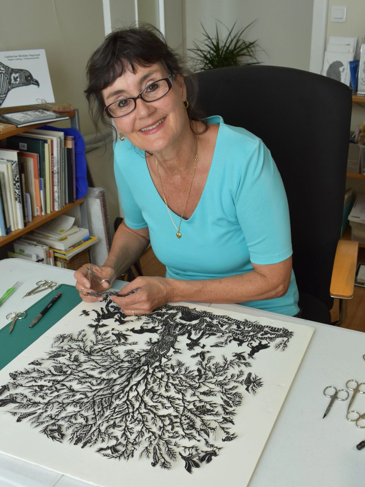 Woman creating detailed paper cut art at a desk, smiling at the camera.
