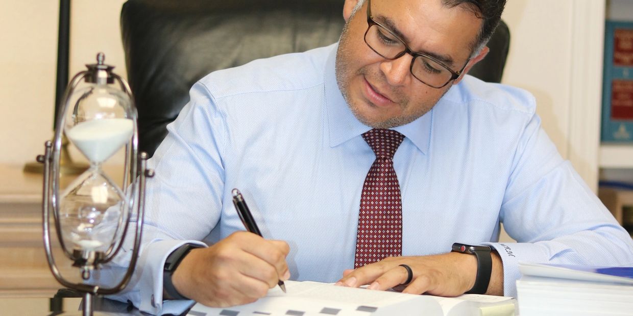 Man in glasses writing at desk with hourglass nearby.