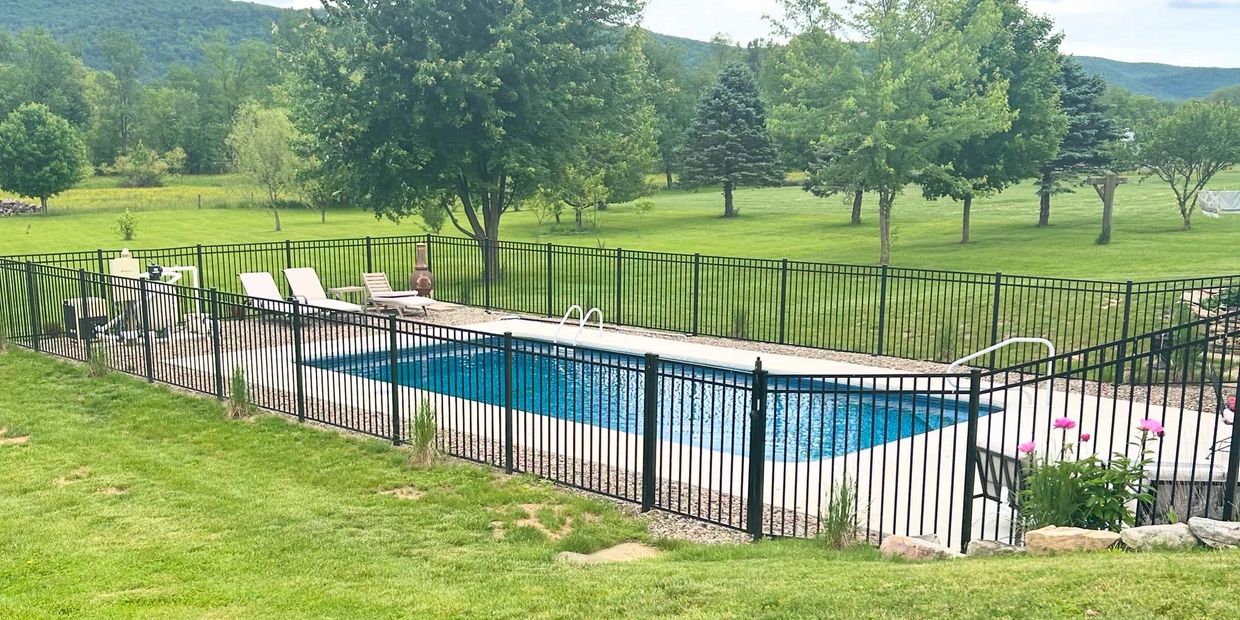A rectangular swimming pool fenced in a lush green backyard with trees and mountains.