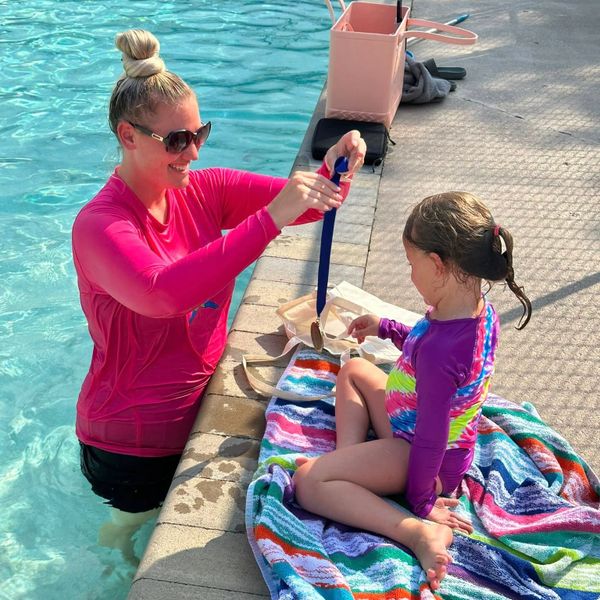 A woman in a pool gives a medal to a girl sitting on a towel by the poolside.