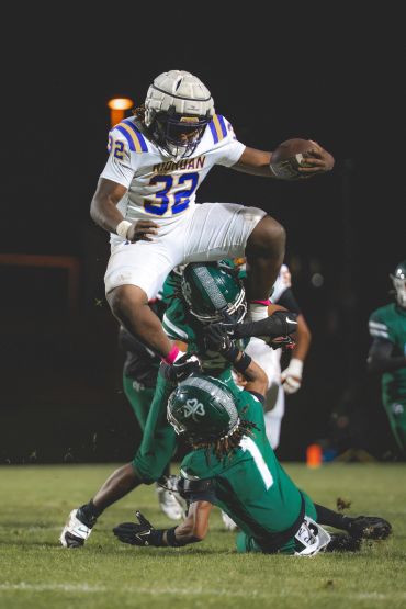Football player in white jersey jumps over two opponents in green.