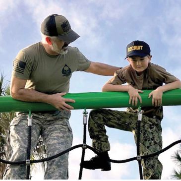 Man helps boy climb obstacle in military attire.