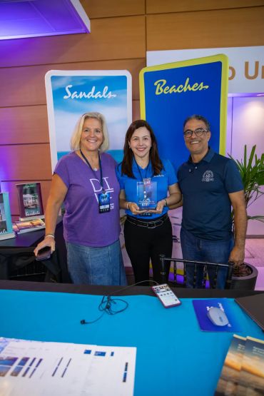 Três pessoas sorridentes em um evento com banners de Sandals e Beaches.