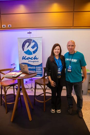 Duas pessoas posando em um evento com banner de Koch Viagens.