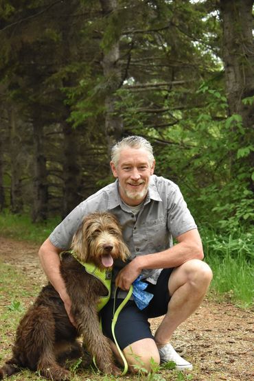 A man kneels with his dog on a forest trail, both smiling happily.