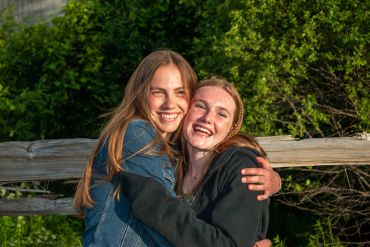 Two young women happily embracing outdoors with greenery in the background.