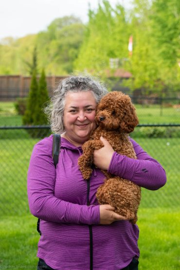 Woman in purple jacket holding a brown curly-haired dog outdoors.