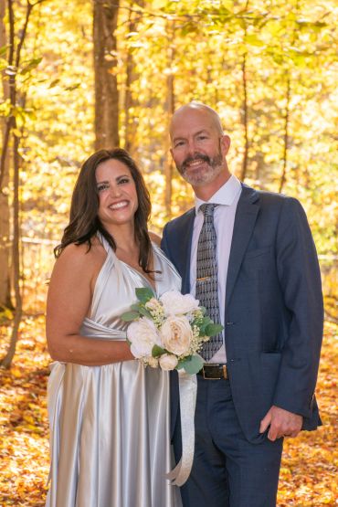 Smiling couple in elegant attire stands in autumn forest holding a bouquet.