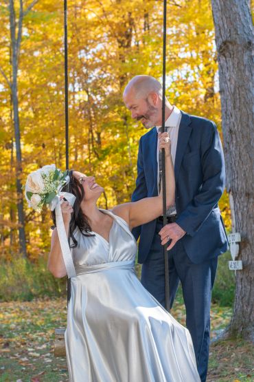 Bride on a swing looks up at groom in a suit amid autumn trees.