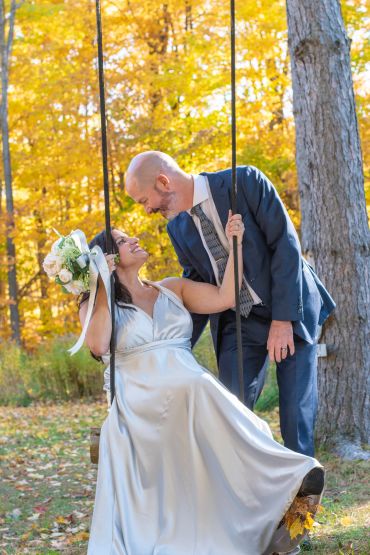 Bride in white dress and groom in suit share a joyful moment on a swing in autumn.