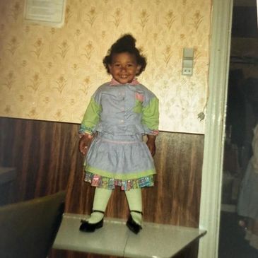 A young girl in colorful clothing stands smiling on a table indoors.
