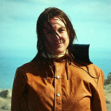 Person wearing a wet brown raincoat with wet hair, smiling outdoors.