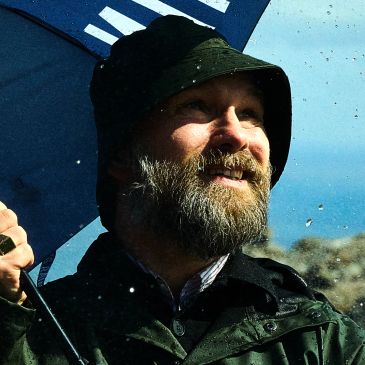Man with beard smiling under a blue umbrella in the rain.