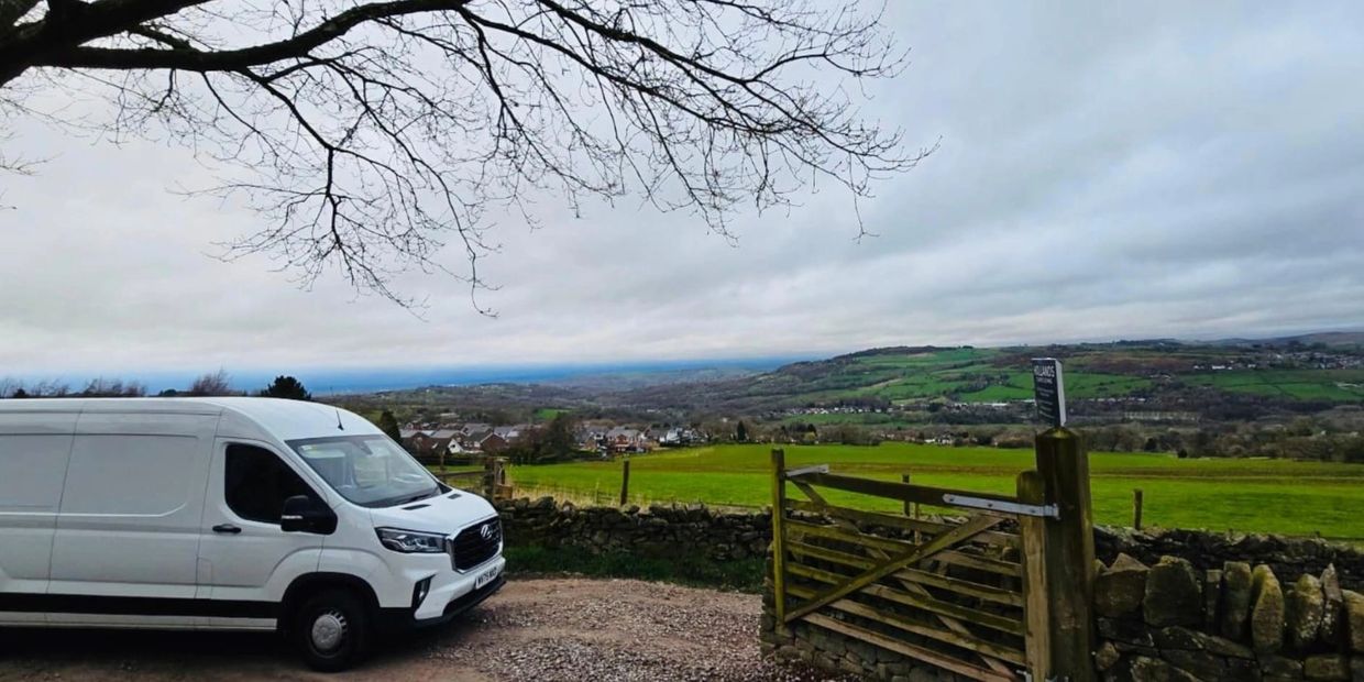 White van parked by a countryside gate with green fields and hills in the background.