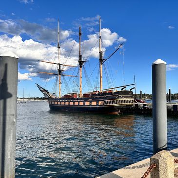 Historic tall ship docked at a sunny marina with blue skies and scattered clouds.