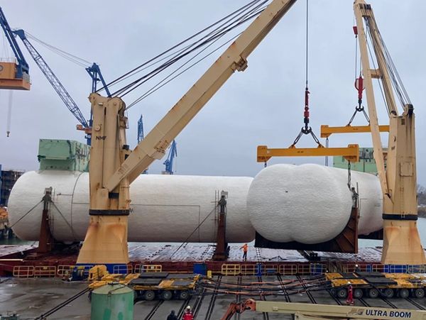 Large industrial tank being lifted by cranes at a dockyard.