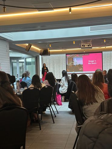 Audience attentively listening to a speaker during a presentation titled 'Stage 1: Be Seen'.