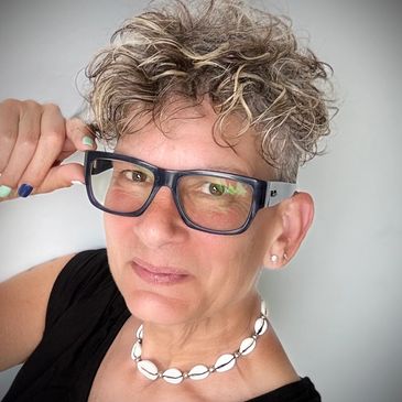 Woman with curly hair, glasses, and shell necklace posing confidently.